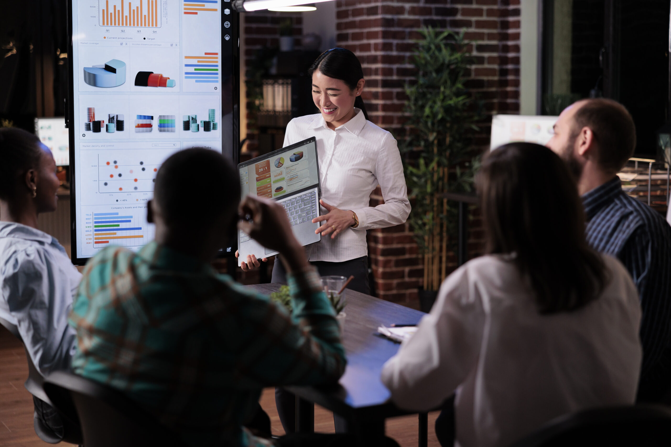 Smiling asian entrepreneur standing in business office presenting turnover charts on laptop screen